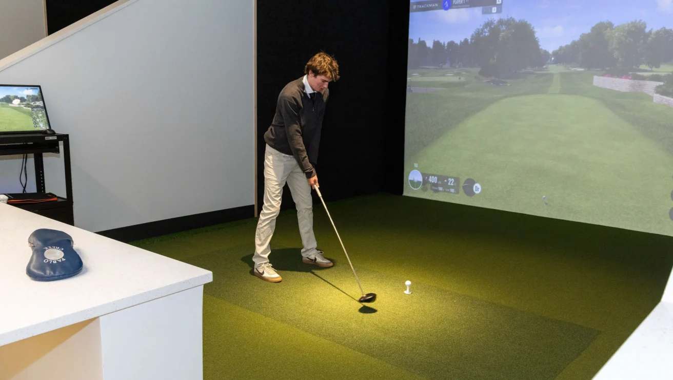 Person playing golf in an indoor simulator, holding a club ready to swing at a golf ball on a mat with a screen displaying a virtual golf course.