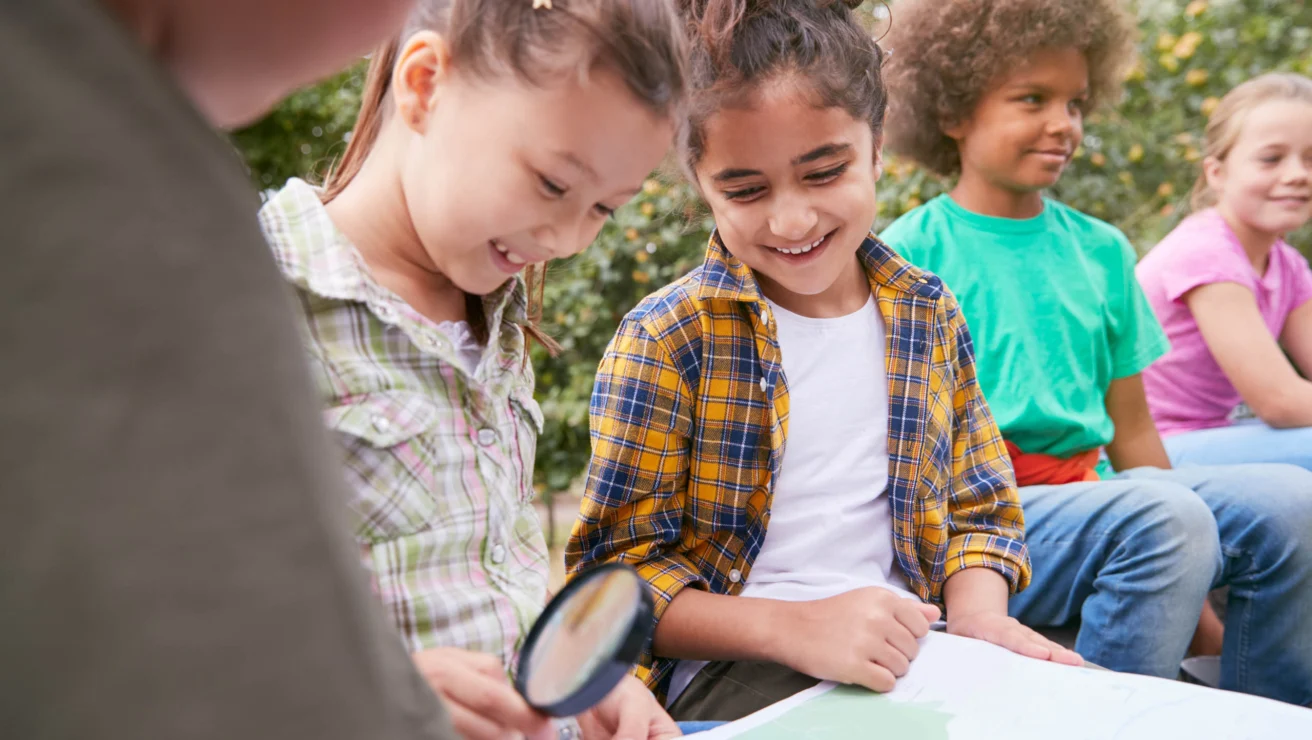Children are sitting outdoors, examining a map with a magnifying glass, while smiling.