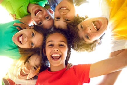Six smiling children in colorful shirts form a circle, looking down at the camera against a bright sky background.
