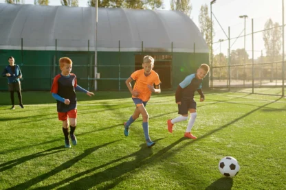 Three boys running on a soccer field towards a ball, with a dome structure and fence in the background on a sunny day.