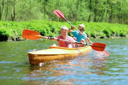 Two people kayaking on a calm river, surrounded by green vegetation. They are paddling with red paddles on a sunny day.