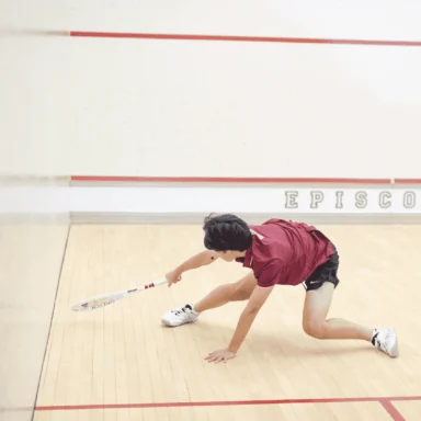 Two squash players in action on a court. One player crouches low to hit the ball, while the other watches, ready to respond. They are wearing athletic clothes and holding rackets.