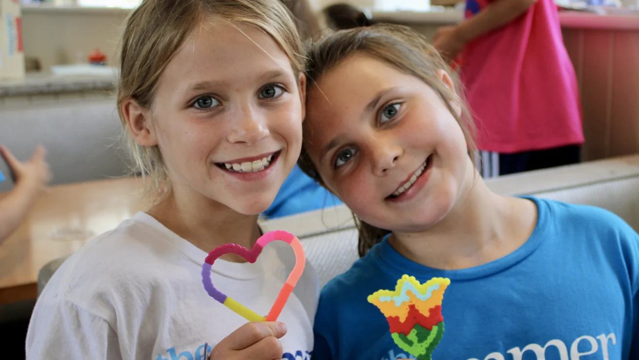 Two smiling girls pose indoors with colorful beaded crafts—one heart-shaped, one flower-shaped. Both wear blue and white shirts from one of the best summer camps in Virginia, creating memories and friendship together.