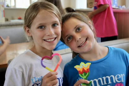 Two smiling girls pose indoors with colorful beaded crafts—one heart-shaped, one flower-shaped. Both wear blue and white shirts from one of the best summer camps in Virginia, creating memories and friendship together.