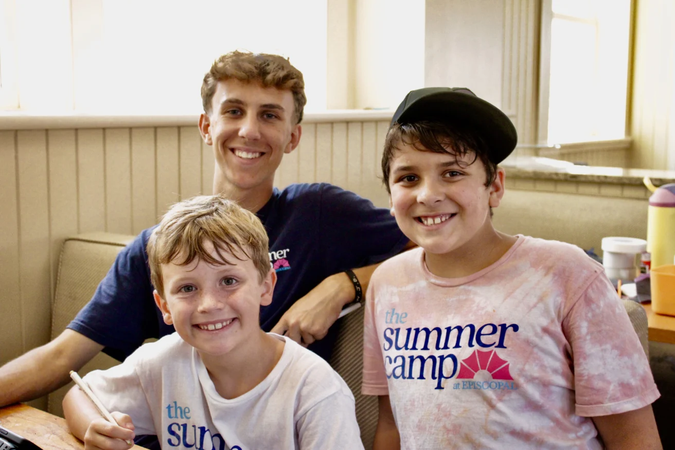 Three smiling boys sit indoors at a table, two wearing “The Summer Camp” t-shirts from one of the best summer camps in Virginia. The older boy has his arm around the younger boys, one holding a pencil as bright daylight streams through the windows.