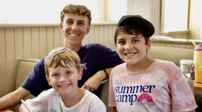 Three smiling boys sit indoors at a table, two wearing “The Summer Camp” t-shirts from one of the best summer camps in Virginia. The older boy has his arm around the younger boys, one holding a pencil as bright daylight streams through the windows.