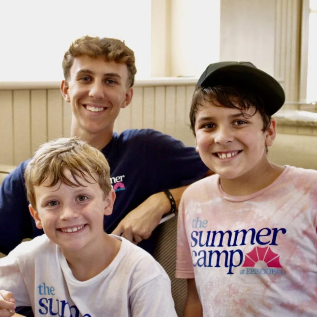 Three smiling boys sit indoors at a table, two wearing “The Summer Camp” t-shirts from one of the best summer camps in Virginia. The older boy has his arm around the younger boys, one holding a pencil as bright daylight streams through the windows.
